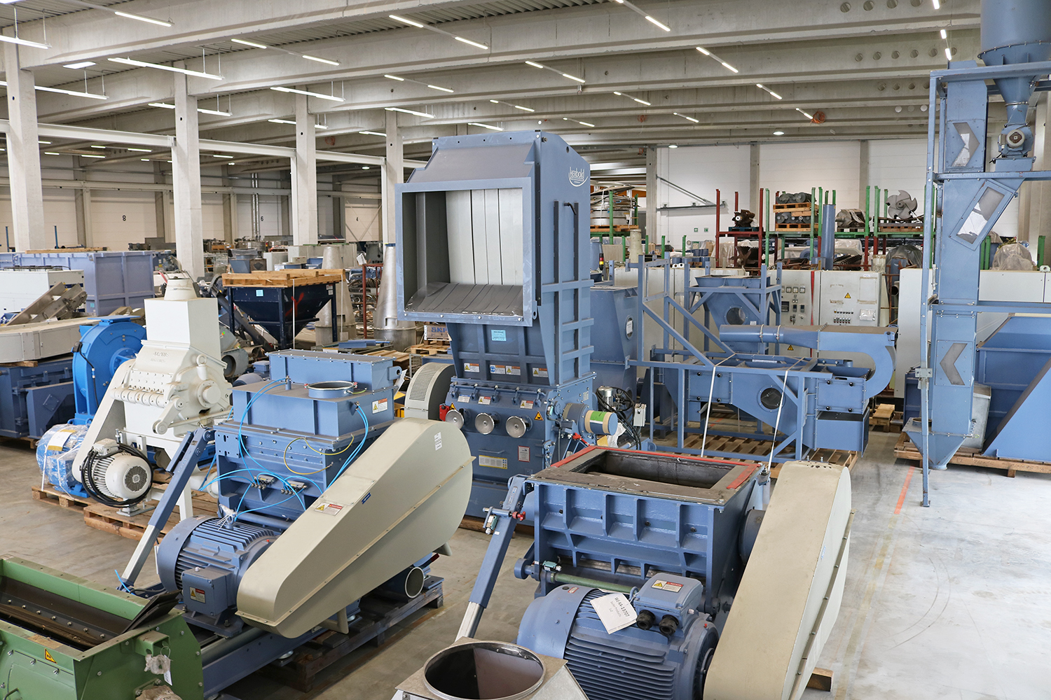 A view inside an industrial warehouse containing various machines for recycling plastics, with individual spare parts on shelves in the background