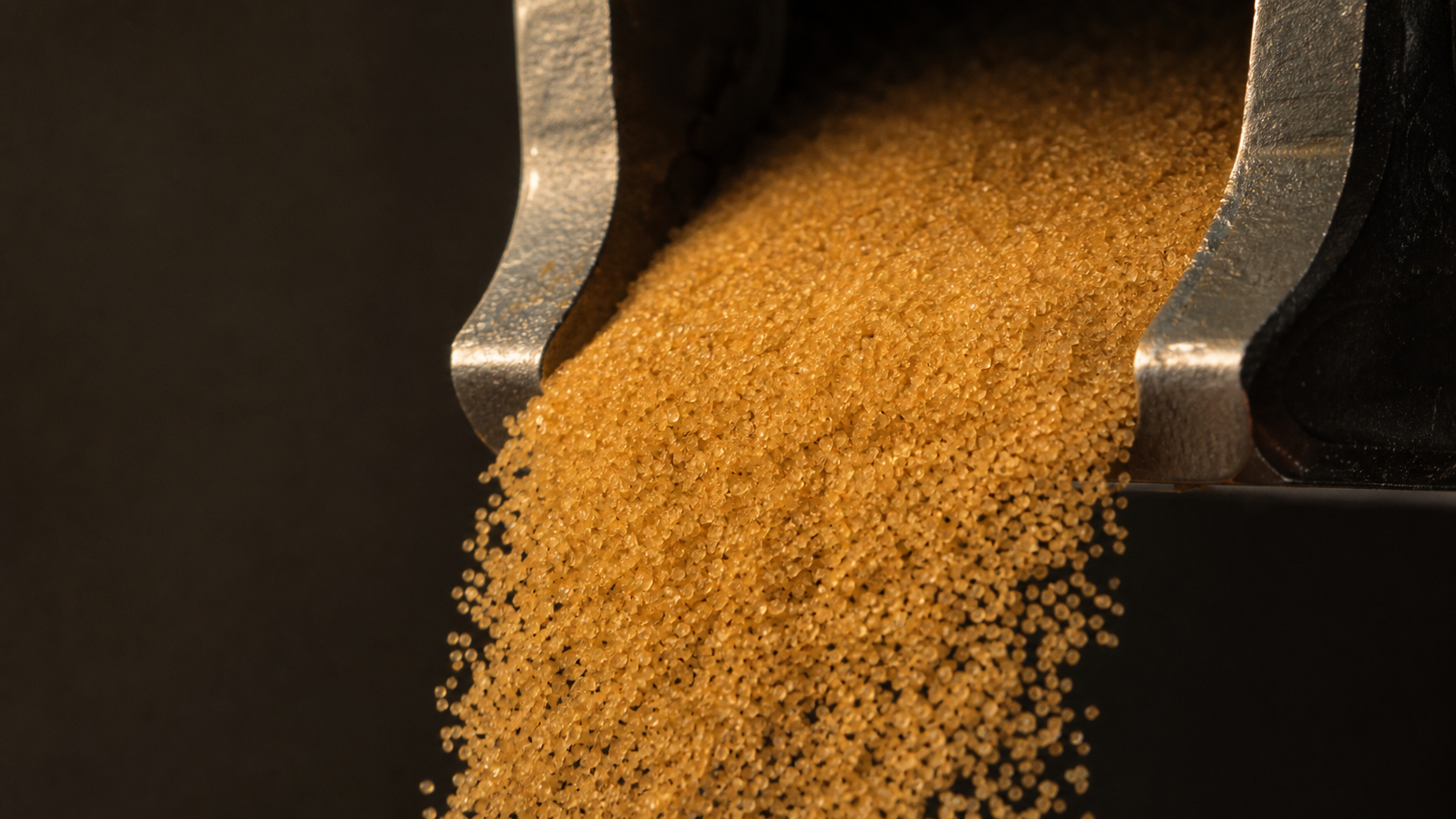Close-up of golden foundry sand flowing through a metallic machine opening, illuminated by warm side-lighting.